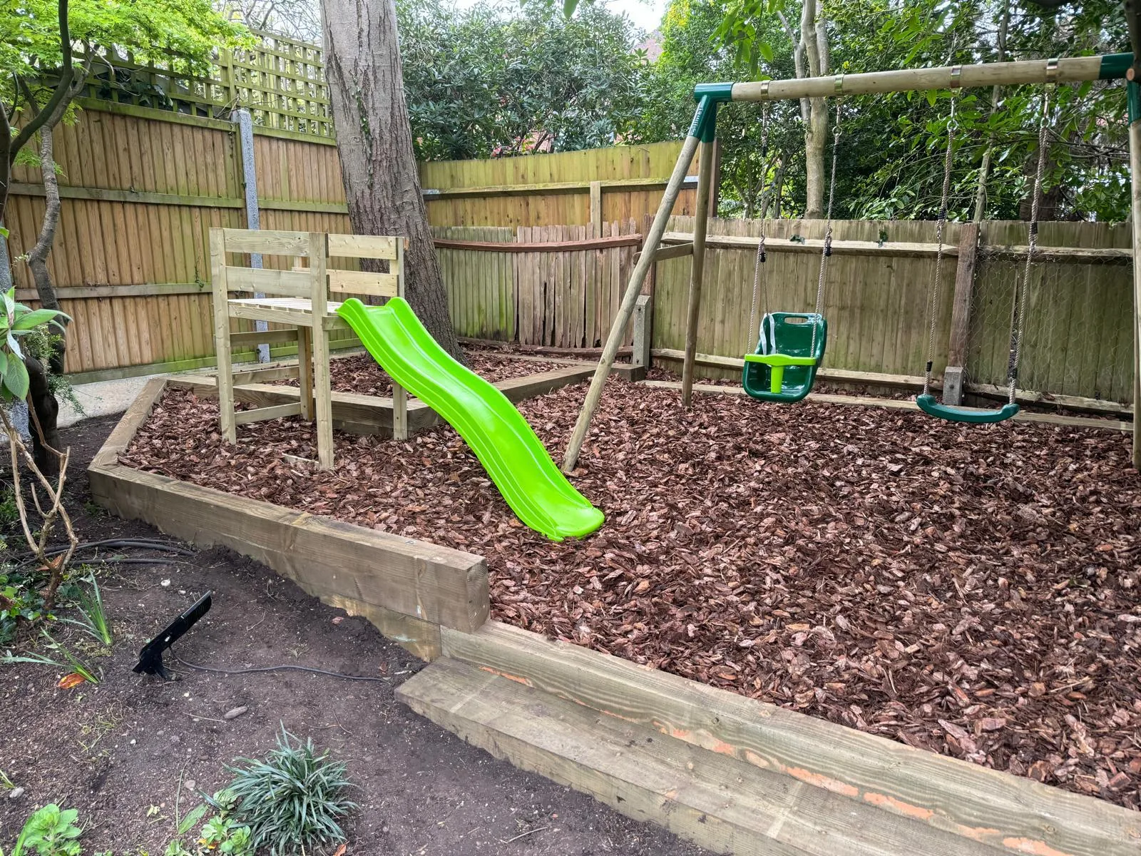 Children's play area with green slide and swing set in a bark-mulched section of garden